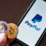 Mão segurando moedas de Ethereum e Bitcoin e um celular com o logo do PayPal ao fundo